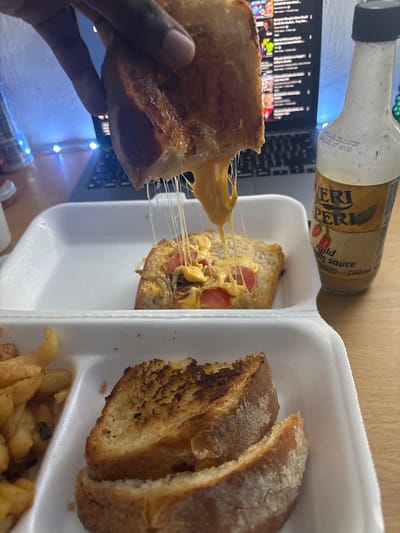 This image depicts a casual indoor dining scene, likely in Port Alfred, South Africa. A dark-skinned hand is shown lifting the top slice of a grilled cheese sandwich, from which numerous strands of melted orange cheese stretch downwards to the remaining bottom half of the sandwich. The sandwich, made with golden-brown toasted bread, features visible melted cheese, red tomato slices, and possibly caramelized onions.

The food is served in a white, multi-compartment foam takeout container. The upper compartment contains the separated halves of the grilled cheese sandwich, while the lower-left compartment holds a portion of golden-brown french fries. Another grilled sandwich half is visible in the lower-right compartment.

On the right side of the frame, a clear glass bottle with a black cap is visible, labeled "VERI PERI mild African sauce, 250 ml." Production dates "20.01.25 L13T25" and "20.01.27 16:30" are faintly visible on the bottle. In the background, an open silver or grey laptop displays what appears to be a streaming service or video platform with multiple thumbnails and text. Blue LED string lights cast a blue glow behind the laptop on the left. The items rest on a light-colored, possibly wooden, tabletop. A small white object is partially visible on the far left of the table.
