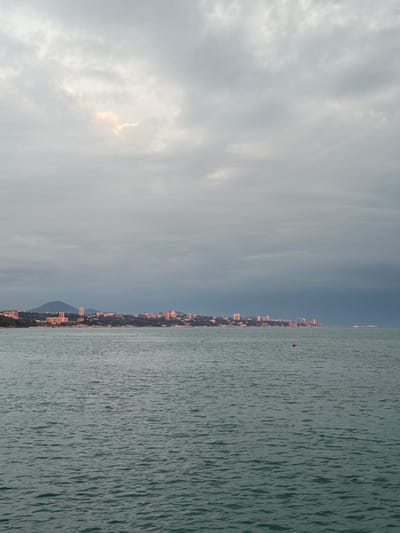 The image depicts a wide, choppy body of water in the foreground, likely the Black Sea, with ripples and waves creating a textured surface. In the middle ground, a distant cityscape stretches horizontally along the coastline. Buildings of various heights, many appearing to be hotels or residential blocks, are clustered together, some illuminated by the soft, diffused light of the overcast sky. Behind the city, rolling hills and mountains form a natural backdrop, their slopes covered in dark green foliage. The sky is dominated by heavy, gray clouds, with a small patch of lighter, pinkish light breaking through near the top left, suggesting either dawn or dusk. There are at least two small, red buoys visible in the water. The overall scene conveys a sense of a coastal city under a cloudy sky. The location is identified as Vardane Microdistrict, Russia.