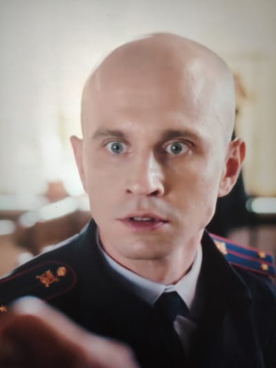 This is a close-up, head-and-shoulders shot of a bald, fair-skinned man wearing a dark military uniform. He has wide, light blue eyes and a pale complexion. His mouth is slightly open, as if he is about to speak or is surprised. The uniform has a dark collar with red and blue trim on the epaulets, and gold insignia on the shoulders. The background is blurred and appears to be an indoor setting with warm lighting, suggesting daytime. The man's expression is one of heightened awareness or concern. The image quality is somewhat grainy, possibly from a video recording. There is no visible text in the image.