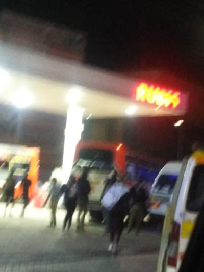 This blurry, low-light photograph depicts a nighttime scene at a Rubis petrol station, likely serving as a transport hub in Oloosirkon/Sholinke ward, Kenya. A prominent red and yellow "RUBIS" sign is visible on the illuminated canopy structure. Beneath the canopy, several indistinct figures of people are present, suggesting movement or waiting activity.
Various vehicles are visible in the scene. A large red and white vehicle, resembling a bus or truck, is positioned towards the center-right background. On the right side of the frame, parts of at least two white vans or minibuses are seen, consistent with public transport vehicles like matatus common in Kenya. The dark sky and bright artificial lighting from the station's canopy confirm the time of day as night. The overall blurriness of the image suggests either motion blur from moving subjects or the photographer, or a low-light capture. No specific emotions or weather details are discernible due to the image quality. The image primarily captures people and vehicles gathered at a petrol station during the night.
