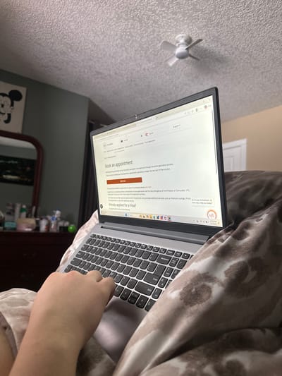 The scene depicts an indoor residential environment, specifically within a room in Yellowhead County, Canada. A light-skinned human right hand is positioned over the keyboard of a silver-colored laptop, indicating active use. The laptop's screen displays a webpage from "VFS Global" titled "Book an appointment," pertaining to visa applications for Austria, identifiable by the Austrian flag in the browser tab. The laptop's taskbar shows the time 12:30 PM and the date 3/11/2020, with an "Ai Assistant" chat window open.

The laptop rests on a patterned blanket or comforter, exhibiting an abstract or animal print in brown and beige tones. In the background, on the left, a dark wooden dresser is partially visible, topped with a large mirror reflecting a darker section of the room. Several small personal items, including bottles and containers, are arranged on the dresser. Above the dresser, on a light green wall, hangs a piece of wall art depicting Mickey Mouse's head in black and white. The upper portion of the image reveals a white, textured (popcorn) ceiling, from which a small, white, three-bladed ceiling fan is suspended and appears to be in motion. A white door or door frame is partially visible in the upper right background.