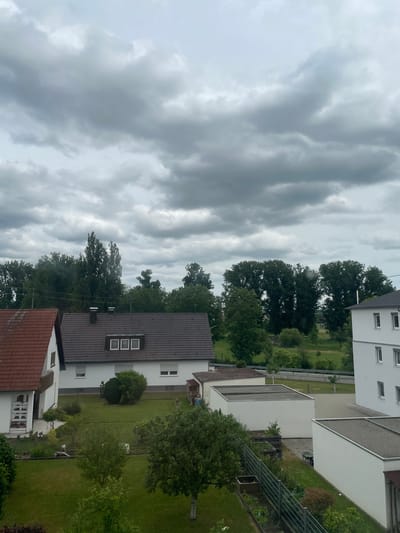 The image, rotated 90 degrees counter-clockwise, depicts an outdoor residential scene under an overcast sky, likely in Günzburg, Germany.

The primary subjects are several houses, lush green trees, and a heavily clouded sky. In the foreground, a house with a dark brown tiled roof features three rectangular dormer windows. To its left, another house with a reddish-brown tiled roof is partially visible. Further back and to the right, a modern white building with a flat, light-colored roof is seen, alongside another white building with multiple windows.

The scene is dominated by greenery, with numerous deciduous trees displaying full, dark green foliage, suggesting late spring or summer. These trees are interspersed between the houses and in the background, where more open grassy areas are visible. Well-maintained green lawns separate the properties, and subtle garden structures, including a metal trellis, can be discerned in some yards.

No people are visible, and no specific activity or event is taking place; the scene is static and quiet.

Notable details include the time of day being daytime, but the weather is heavily overcast, with dense, dark grey clouds dominating the sky, casting a muted light over the landscape. The architecture, characterized by tiled roofs and neat suburban planning, is consistent with typical residential areas in Germany. The overall mood conveyed is calm and serene, perhaps a little subdued due to the heavy cloud cover. There is no visible text in the image.