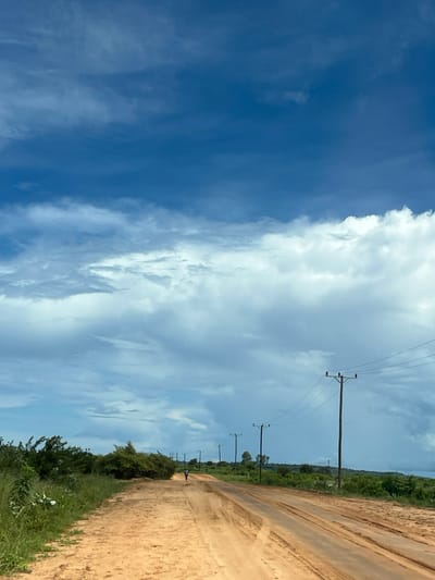 The image depicts a rural or semi-rural scene in Muxara, Mozambique, under a dynamic sky featuring a mix of deep blue and extensive white and grey cloud formations. A wide, unpaved reddish-brown dirt road dominates the lower half of the frame, showing distinct vehicle tracks extending into the distance and receding towards the upper left. Along the right side of the road, a series of wooden utility poles with power lines are visible, also stretching into the background. Lush green vegetation, comprising bushes and trees, borders both sides of the road. A lone individual is observed walking away from the viewer in the far distance on the dirt road, positioned centrally. The overall environment appears natural and undeveloped, with signs of human infrastructure and passage.