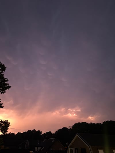 A low-angle, vertical shot captures a dramatic twilight sky over the city of Putten, Netherlands. The sky transitions from a deep, dark purple at the top to a vibrant pink and orange glow near the horizon. Unusual, bulbous cloud formations are visible in the upper portion of the sky. Jagged bolts of lightning streak across the lower half of the sky, illuminating the pink and orange clouds from within. Silhouetted against the colorful sky are the dark shapes of trees and the rooftops of houses. The overall impression is one of a powerful and colorful storm approaching or passing.