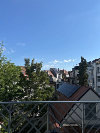 The image captures a view of rooftops in the city of Saint-Gilles - Sint-Gillis, Belgium, under a clear blue sky with scattered white clouds. In the foreground, a grey metal railing with a diamond pattern divides the view. Behind the railing, lush green trees with dense foliage are visible, interspersed with the roofs of buildings. Several buildings feature terracotta-tiled roofs, some of which are equipped with solar panels. The architecture suggests a residential urban setting. The lighting indicates it is daytime, likely morning or afternoon. No people or animals are present in the frame.