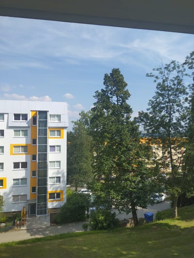This is a medium-shot, eye-level exterior photograph taken on a sunny day. The scene is an urban residential area, likely in Chemnitz, Germany. In the foreground, a sloped grassy area with dappled sunlight suggests an outdoor recreational space. Behind this, a paved walkway leads to a modern apartment building with a white facade accented by yellow geometric shapes and a prominent glass elevator shaft. The building features numerous windows, some with white frames and others with purple. Further in the background, other residential buildings with orange facades are visible, peeking through lush green trees. A street with parked cars, including a white hatchback, runs alongside the buildings, and a blue trash bin is positioned on the side of the road. The sky is a clear blue with scattered white wispy clouds, indicating pleasant weather. The time of day appears to be late morning or early afternoon due to the bright sunlight and shadows. There are no people visible in the frame, and the overall mood is tranquil and ordinary. No text is visible in the image.