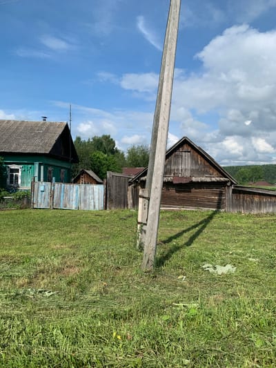 This is a full-frame, eye-level, outdoor, daylight, medium shot of a rural scene. The background consists of a bright blue sky with scattered white clouds. In the mid-ground, there are several wooden structures, including a green wooden house with a grey roof on the left, and a rustic wooden barn-like building with a dark wood exterior and a grey roof on the right. A weathered blue and white wooden fence separates the green house from the rest of the yard. There are also several trees and vegetation in the background. In the foreground, a large, rough wooden utility pole stands vertically, casting a long shadow across the freshly cut green grass. The ground is covered in green grass, with some areas showing cut grass clippings. The scene appears to be a residential area in the countryside. The location is identified as Barsai, Russia.