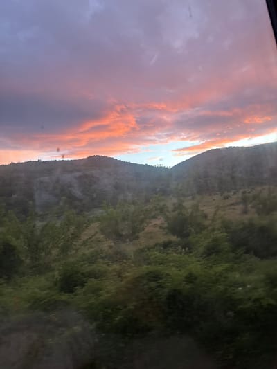 The image shows a landscape at dusk or dawn. On the left side, there's a sky with purplish-gray clouds transitioning to orange hues near the horizon, suggesting either sunrise or sunset. To the right, a hillside covered in vegetation, primarily green with varying shades, appears. The landscape is divided by the ridgeline of the hill, with the sky on one side and the vegetated slope on the other. There are no people visible, and the environment appears natural. The image seems to have been taken from an elevated position, perhaps from a moving vehicle. Given the prompt's mention of Binkos, Bulgaria, it can be inferred this is a landscape near that city. However, no specific details tie the scene directly to Binkos, Bulgaria, as it could be any hillside landscape.