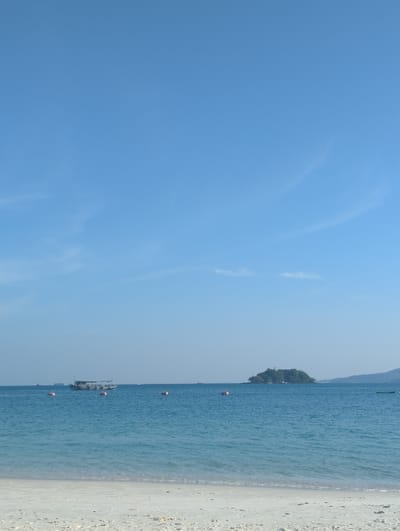 The image displays a coastal scene within Preah Sihanouk, Cambodia, characterized by a clear sky, calm ocean, and a sandy beach. The foreground features a strip of light-colored sand. The mid-ground is dominated by turquoise-blue ocean water with gentle ripples. A dark-hulled boat, approximately medium-sized, with a cabin structure, is positioned in the mid-ground, slightly to the left of center. Four orange-red spherical buoys are scattered across the water. In the mid-to-far distance on the right, a small, densely vegetated island is visible, backed by a more distant, hazy landmass on the horizon. A smaller, dark boat is faintly visible near the island on the far right. The sky overhead is a clear, bright blue with minimal, wispy white clouds present in the lower quadrants. No individuals are discernable on the beach or within the visible boats.