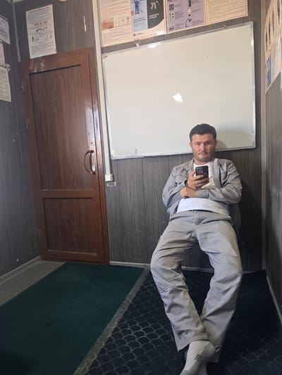 A man in a grey work uniform is sitting on the floor indoors, holding a smartphone and looking at the camera. He is in a narrow hallway with a wooden door to his left and a whiteboard on the wall behind him. The walls are paneled with dark grey wood-effect material. There are posters on the wall above the whiteboard. A green mat is on the floor in front of the door, and a patterned carpet is to the right of the mat. The environment suggests a utility area or a break room in an industrial or institutional setting. The man's posture and expression are relaxed. The location is identified as Jararyk, Uzbekistan.