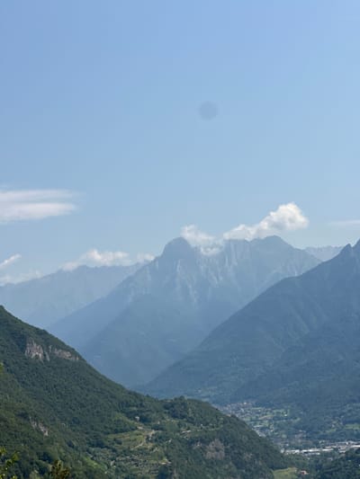 This is a distant landscape photograph taken from a high vantage point, likely on a hillside overlooking a valley. The scene is dominated by layers of mountains under a clear, pale blue sky. In the foreground, a steep, tree-covered slope descends into the valley. The trees are lush and green, with visible terracing on some parts of the slope, suggesting agricultural use. Further into the valley, a small town or village is nestled among the trees. Buildings are visible, with hints of roads and infrastructure. Beyond the town, more mountain ranges recede into the distance, becoming progressively lighter and hazier due to atmospheric perspective. Wisps of white clouds are present in the sky and around some of the higher mountain peaks. There are no people or animals visible in the frame. The overall impression is one of a vast, natural landscape with human settlement integrated into it. The location is identified as being in or near Malegno, Italy, which aligns with the mountainous terrain and the style of architecture visible in the village.
