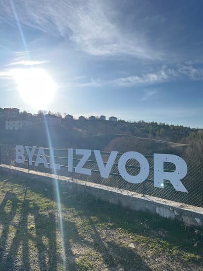 The image captures an outdoor scene in Byal Izvor, Bulgaria, under a clear blue sky with sparse, wispy white clouds. A prominent white sign spelling "BYAL IZVOR" in large, uppercase, sans-serif letters is mounted on a chain-link fence, positioned horizontally across the midground. The fence features a concrete base. Strong sunlight emanates from the upper left, creating a significant lens flare and casting long shadows across the patchy green and brown grass in the foreground. In the background, a hillside rises, featuring several residential buildings, predominantly on the left side, interspersed with bare deciduous trees and denser evergreen foliage further to the right. No individuals are visible in the scene.
