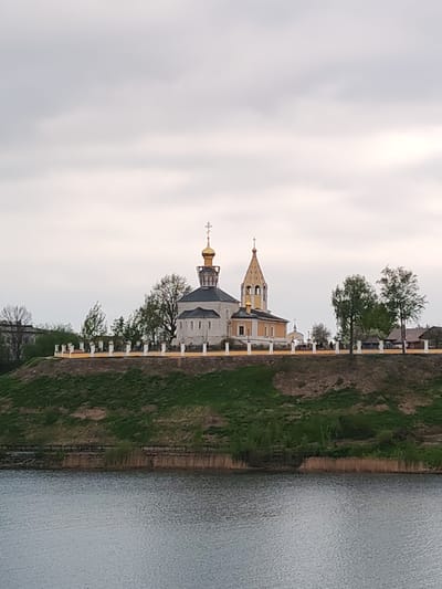 The image depicts a church atop a grassy hill in Gorodnya, Russia, as viewed across a body of water. The church features a gold dome and a bell tower, surrounded by a white fence. Trees are interspersed along the hilltop. The sky is overcast. Reeds line the water's edge.