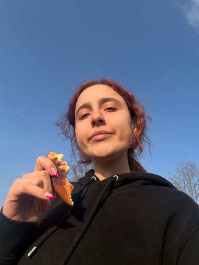 A young female, with reddish-brown hair pulled back, is depicted outdoors in Domgermain, France, on a clear day with a bright blue sky. She wears a black hooded sweatshirt with visible silver eyelets for the drawstrings. Her left hand, featuring manicured nails with a white base and red wavy designs, holds a partially consumed piece of golden-brown flatbread with a slightly charred crust. She is looking directly at the camera with a neutral to slightly amused expression. In the background, to the right, bare tree branches are visible against the sky. The lighting suggests natural sunlight from the left, casting subtle shadows on her face.