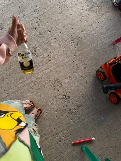 The image captures a close-up, high-angle view of a concrete ground surface. In the upper left quadrant, a hand with pale skin and light yellow nail polish is holding a clear glass bottle of Corona Extra beer, with the label partially visible. Below this, a person's legs are visible, wearing light blue distressed jeans and pink Birkenstock sandals. The person is also wearing a brightly colored garment with a cartoonish yellow character, possibly part of a shirt or jacket. To the right of the frame, an orange toy lawnmower with black accents and orange wheels is present. A person's lower leg, clad in a dark boot, is positioned next to the toy. In the lower right quadrant, a red cylindrical object with white caps on both ends lies on the concrete. Several green plastic objects, possibly parts of a toy or a game, are also visible in the lower right corner. The overall scene suggests an informal outdoor setting, possibly a backyard or patio. There is no identifiable information to place this scene in Пепеляева, Russia.