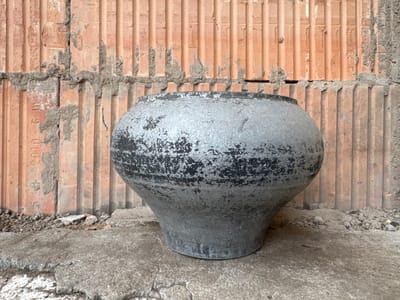 The image shows a large, weathered, grey cast-iron pot resting on a concrete surface in front of a wall of unfinished, orange-colored porous bricks. The bricks are laid in a vertical pattern with visible mortar lines and some crumbling edges. The concrete surface is rough and textured, with debris and smaller pieces of brick scattered around the base of the pot. The pot itself is bulbous, with a wide opening and a narrower base. Its surface is uneven and shows signs of age and wear, with patches of black and grey discoloration. The background wall features some faint markings, possibly dates or manufacturer stamps, including "2010". The overall impression is one of an outdoor, possibly construction or storage area, with the pot appearing to be an object of some age and utility, abandoned or temporarily placed in this industrial-looking setting. The scene is devoid of any people or animals. The lighting is even, suggesting an overcast day or diffused light.