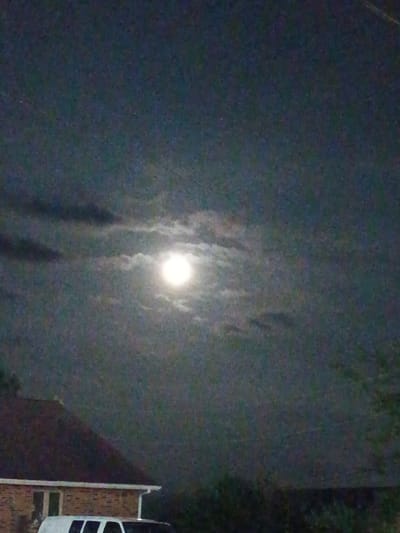 The image captures a nighttime scene with a full moon prominently featured in the sky, partially obscured by clouds. Below the sky, the roofline of a brick building is visible, with a white vehicle parked in front of it. The environment appears to be suburban, with hints of trees and vegetation. The location is identified as Nicholasville, United States of America.