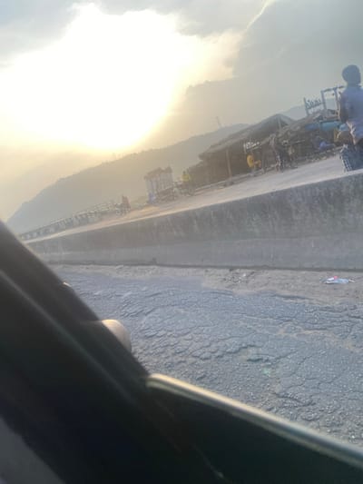 The image, rotated 90 degrees clockwise, captures a vibrant outdoor scene, likely from the perspective of someone inside a vehicle. The composition is split diagonally by a long concrete barrier, separating a busy roadside market on one side from a textured road surface on the other.

On the left side of the image, the sky is dominated by an intensely bright, overexposed light source, almost certainly the sun, which appears low on the horizon, suggesting either sunrise or sunset. This powerful glare creates a lens flare and washes out a significant portion of the sky in brilliant white, transitioning to hazy light yellow and orange hues around its periphery. Scattered wispy clouds are visible in the sky. In the far distance, partially obscured by the haze and light, a range of mountains or large hills stretches across the background. A tall, thin structure, possibly a communication tower or an electric pylon, can be seen standing on one of the distant peaks.

Parallel to the bright sky and mountains, to the right of the concrete barrier, lies an active roadside area. This section features what appears to be an informal market or a row of small, makeshift shops. Various structures, likely temporary shelters made of tarpaulins and other materials, are lined up, seemingly cluttered with goods. Several people are visible in this area, engaged in activity. One individual, wearing a light blue top and a dark head covering, is prominently featured near the upper part of this market section. Other figures are seen further down among the stalls. Bicycles and small, loaded carts or rickshaws are also present, indicating local commerce and transportation.

The right side of the image reveals the immediate foreground: a dark, heavily textured road surface, likely asphalt or worn concrete. This surface is characterized by numerous cracks, undulations, and small irregularities. A discarded plastic bottle, light in color, lies on this ground near the concrete barrier. In the extreme bottom-right corner, a dark, curved section of what appears to be a vehicle's interior (possibly a window frame or door panel) is visible, confirming the vantage point from inside a car or similar vehicle.

The overall atmosphere is bright and hazy, with the warm light suggesting either early morning or late afternoon. There is no discernible text visible in the image.