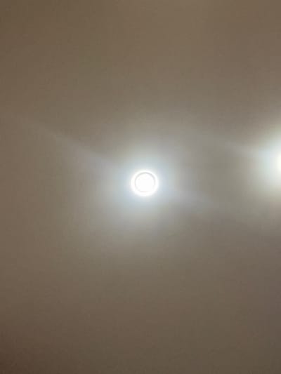 The image depicts a close-up, overhead view of a beige, textured ceiling. Two circular, recessed lights are visible. The light on the left is centered and emitting a bright white glow, with a distinct circular halo around it. The light on the right is positioned towards the upper right corner of the frame and is also emitting a bright white light, with a less pronounced halo. The texture of the ceiling appears somewhat grainy. There are no people, objects, or actions evident in the image, other than the illuminated ceiling fixtures. The context of Bugayevka, Russia, is not visually represented in the provided image.