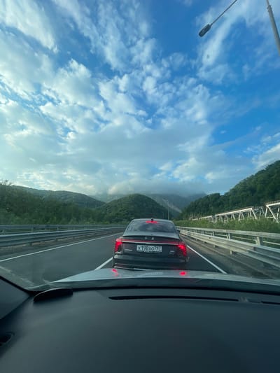 The image shows a view from inside a car driving on a highway. In front, another car is visible with a license plate reading "X198XO797", indicating the location is likely in Russia. The landscape features green mountains and a cloudy sky. The presence of guardrails and a bridge structure suggests the highway is elevated. The location could potentially be near the city of Monastyr, Russia, given the context.