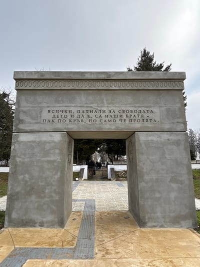 A concrete archway monument stands under an overcast sky in Shumentsi, Bulgaria. The structure comprises two thick, untextured grey concrete pillars supporting a horizontal lintel. The lintel's upper edge features an engraved decorative band of repeating oval motifs. Centered below this, on the front face of the lintel, is an inscription in Cyrillic: "ВСИЧКИ, ПАДНАЛИ ЗА СВОБОДАТА - ДЕТО И ДА Е, СА НАШИ БРАТЯ - ПАК ПО КРЪВ, НО САМО ЧЕ ПРОЛЯТА." This translates to: "ALL WHO FELL FOR FREEDOM - WHEREVER IT MAY BE, ARE OUR BROTHERS - AGAIN BY BLOOD, BUT ONLY THAT WHICH WAS SHED." On the inner vertical faces of the pillars within the archway, circular etched designs or emblems are visible, one on the left pillar appearing to contain numbers, possibly "19XX," within a wreath border. The ground leading to and through the archway is paved. The immediate approach features reddish-brown stamped concrete with a central grey tactile paving strip containing raised parallel ridges. Small circular metal fixtures are embedded in the reddish paving. Beyond the archway, the path continues with lighter grey square pavers, leading to a dark metal gate. Several individuals, dressed in dark outerwear, are visible beyond the gate, appearing to walk or gather. Low white walls and grey stone posts flank the path beyond the archway. The environment includes sparse green grass and a mix of bare deciduous and evergreen conifer trees under the grey, uniformly clouded sky.