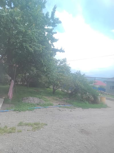 An outdoor scene shows a gravel driveway in the foreground. Behind the driveway, a grassy area is visible with several trees. A pink cloth hangs from a metal pole on the left side of the frame. In the middle ground, a pile of dirt is present, and a blue hose runs along the edge of the grass. Further back, a residential building with a red roof is partially visible, along with another building to its right. A yellow barrel is situated near the buildings. The background consists of a cloudy sky and distant hills. The image was taken in the city of Esik, Kazakhstan.