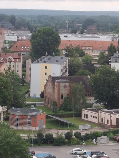 An aerial view of a city shows a densely built environment with a mix of residential and industrial buildings. The foreground features a circular, red brick structure with horizontal louvered windows, possibly a utility building, adjacent to a longer, low-rise cream-colored building with multiple windows. Beyond these, a mid-rise building with a white and yellow facade stands out. In the background, larger, multi-story buildings with orange tiled roofs and numerous windows dominate the skyline, interspersed with mature green trees. The overall impression is of an urban landscape under an overcast sky. This scene is located in the city of Pila, Poland, specifically in the area of the former Pila military airfield, which has since been redeveloped for civilian purposes.