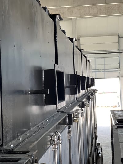 The image shows a close-up, low-angle, side view of a row of large, black, industrial metal structures with visible boxes protruding from them. The structures are mounted on a silver track or rail system with visible bolts and clamps. The background reveals a bright, white, industrial building interior with a large open doorway or garage door. The floor appears to be concrete. The scene is located in Modřice, Czechia. There are no people visible in the image.
