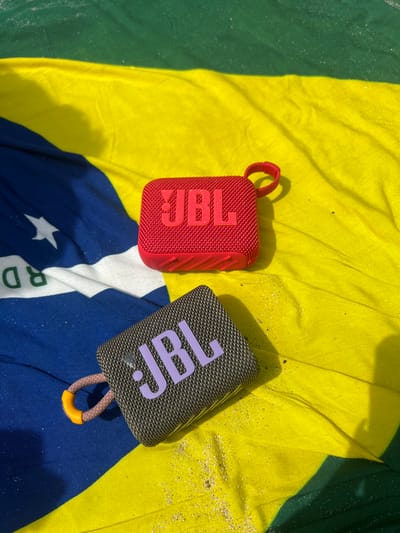The image shows two JBL portable Bluetooth speakers lying on a beach towel. The speakers are both in focus, with the red speaker on top and the black speaker on the bottom. The beach towel is folded, with the green and yellow portions visible. The red speaker has a black strap attached to it and a light pink "JBL" logo. The black speaker has an orange strap attached to it and a purple "JBL" logo. The speakers are in the city of Santa Fe, Costa Rica, at the coordinates latitude: 9.642, longitude: -85.168.