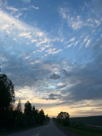 A landscape view in Zykovo, Russia presents a divided sky: the left portion exhibits a clear, light blue hue with scattered, wispy clouds, while the right portion features a dense layer of grey and white clouds, illuminated by a faint golden light, possibly from the setting sun.
Below the sky, a dark treeline stretches along the right side of the frame. To the right of the treeline, a paved road curves into the distance. A hint of green vegetation is visible on the far right edge, suggesting a field or hillside.