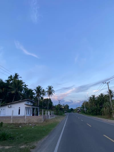 The image depicts a rural road in Tanjongkeumala, Indonesia, during twilight. A single-lane asphalt road stretches into the distance, bordered by lush green vegetation and palm trees. On the left side of the road, a modern white house with a dark roof and a covered porch is visible. A low fence made of concrete and brick encloses the property. Utility poles with power lines are present along the road, indicating an established infrastructure. The sky is a clear blue with streaks of wispy white and pink clouds, characteristic of the sunset or sunrise. The scene appears peaceful and tranquil, with no people or vehicles visible.