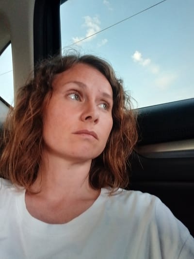 A woman is seen from the chest up, her head turned to her right. She is wearing a white t-shirt and has shoulder-length, wavy brown hair. The background shows a light blue sky with a few white clouds and what appears to be a power line. The setting seems to be inside a vehicle, with part of a car's interior visible on the right side of the image. The woman is looking out the window, her expression thoughtful. The lighting suggests it might be late afternoon or early evening. The image was captured in the city of Yalgyz-Narat, Russia.