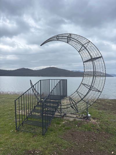 A large, skeletal metal crescent moon-shaped decorative structure, approximately several meters in height, stands on a grassy embankment overlooking a body of water. The crescent, oriented with its concave curve facing left, is constructed from a dark-colored metal frame and covered with numerous unlit white or clear light elements. A black metal staircase with matching handrails is attached to the left side of the crescent, leading to a small, elevated square platform positioned within the moon's curve. The immediate foreground features green grass mixed with patches of exposed dark soil and sparse small purple flora. Beyond this, a calm, rippled body of water, identified as the Koprinka Reservoir, extends across the midground. Distant, undulating hills or mountains, covered with bare trees, form the horizon. The sky is uniformly overcast grey. No individuals, actions, or interactions are present in the scene. The location is at Koprinka Reservoir, near the town of Pavel Banya, Bulgaria.