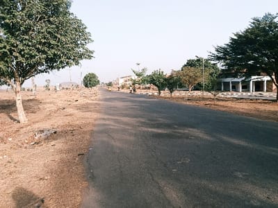 The image displays an unidentified asphalt road within Rafin Pa, Nigeria, under a clear, bright sky, suggesting daytime with long shadows indicating morning or late afternoon. The road, appearing to be two lanes wide, extends into the distance. On the left, the immediate foreground consists of dry, reddish-brown earth with sparse, short vegetation. A mature tree with green foliage stands prominently on the left, casting a shadow. Further left, utility poles are visible, and distant hills or mountains rise on the horizon.
On the right side of the road, a strip of dry ground parallels the asphalt, bordered by a series of white and black painted curb stones. A row of smaller, leafy trees lines this side of the road. Beyond these trees, a multi-story building with a facade featuring several white pillars and dark, open sections, possibly entrances or windows, is prominent. This structure appears to be either under construction or disused.
In the mid-ground, on the right side of the road, two individuals are walking away from the viewer. Further down the road, also on the right, a yellow motorized tricycle, commonly known as a keke Napep, is visible. Streetlights are sparsely distributed along the road's edges. The ground on both sides of the road is dry, showing signs of arid conditions.