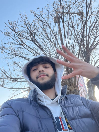 A male individual is depicted in a close-up outdoor selfie, situated in the city of Qudsaiya, Syria. The subject, appearing to be in his late teens or early twenties, has dark, curly hair and a beard. He is wearing a white hooded sweatshirt with the hood pulled over his head, overlaid by a dark blue or black puffy winter jacket. A colorful, geometric pattern is visible on the front of his white sweatshirt. The subject is looking directly into the camera with a neutral expression. His left hand is raised to the left of his face, palm facing outwards, with four fingers extended and the thumb bent, forming a numerical "four" gesture. The background consists of a clear, light blue sky and several bare deciduous trees with visible branches and small, round, yellowish seed pods. A utility pole or lamppost is partially obscured by the tree branches on the right side of the frame.