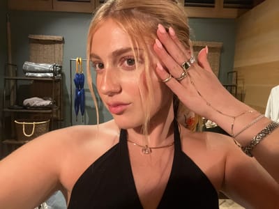 A woman with light blonde hair is facing the camera and holding her hand near her face. She is wearing a black top and gold jewelry around her neck, wrist, and fingers. The background is a room with a shelf containing baskets, towels, and umbrellas. The location is not discernible as Livingstone, Zambia based on visual elements in the image alone.