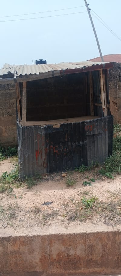 The image captures an unoccupied, open-fronted roadside stall or kiosk in Ado-Ekiti, Nigeria. The structure's lower exterior is composed of dark, weathered corrugated metal sheeting, exhibiting dents, creases, and sporadic reddish-orange paint marks. The upper frame is constructed from rough wooden planks, supporting a rusted and worn corrugated metal roof. An empty wooden shelf or counter is visible within the stall's interior. The background immediately behind the stall features a cinder block wall. The ground in the foreground consists of dry, light brown soil mixed with small stones and sparse green vegetation. A weathered concrete curb runs across the bottom of the frame. In the distant background, under a pale sky, a utility pole with power lines is visible, along with a partial view of another building's reddish-brown corrugated roof and a black water storage tank situated on a separate rooftop. No individuals, merchandise, or vehicles are present in the scene.