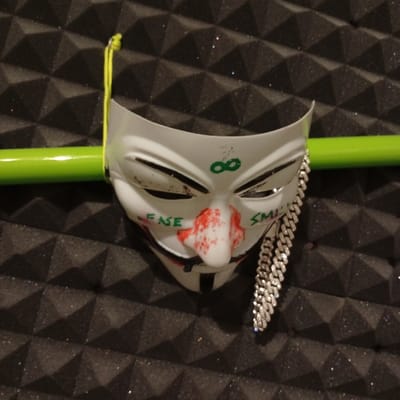 A white plastic mask depicting the face of the character "V" from the film "V for Vendetta" is lying on a surface covered in dark grey acoustic foam tiles. The mask has green lettering on it, reading "FASE" on the left side of the nose and "SMIL" on the right. The mask has a red-painted nose and features a chain-like accessory hanging from its right side. The mask is positioned in a way that it looks like it is wearing the chain. The mask is resting on a green pipe that has a yellow string attached to its upper left side. The location is Biedenkopf, Germany, which is a town in the Lahn-Dill-Kreis district in the state of Hesse, Germany. The coordinates are: latitude: 50.914, longitude: 8.465.