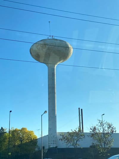 The image depicts a distinctive water tower in Don Torcuato, Argentina. The tower is painted grey. Powerlines are visible in the foreground against a clear blue sky. Street lamps and trees surround the base of the tower. A building abuts the support structure of the tower.