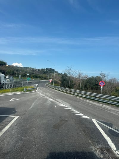 The image depicts a static view of a road junction or on/off-ramp system, consistent with a highway exit in the vicinity of Tortoreto, Italy, indicated by a partially visible green directional sign showing "ANCONA."

The scene is under a clear, bright blue sky with minimal wispy clouds. The terrain features vegetated hills in the background, populated by both evergreen and bare deciduous trees.

In the foreground, a dark asphalt road surface is visible. On the left, a white solid line delineates a lane with a large white triangular arrow pointing left. Towards the center and right, the main road curves gently to the right and ascends. This curving road segment is marked with a continuous line of white triangular "shark's teeth" markings, indicating a yield requirement for traffic entering or merging.

Silver-colored metal guardrails line both sides of the curving road and a separate road section to the far left. Several tall, slender street lamp poles with downward-curving fixtures are positioned along the roadside.

Multiple traffic signs are present:
*   On the left, a green rectangular sign with white text, partially obscured, clearly shows "ANCONA."
*   On the right, near the start of the curve, a red triangular yield sign (Italian "Dare Precedenza") is affixed to a pole.
*   Further along the right side, behind the guardrail, a circular red sign with a blue center and a red diagonal cross (Italian "Divieto di sosta e fermata" – No stopping or parking) is mounted, with a supplementary white rectangular sign below it featuring a downward-pointing blue arrow.

No people are visible. Partial views of multiple white truck cabs are discernable in the far left background, suggesting parked vehicles. Patches of yellow flowers are visible in the grassy median strip on the left.