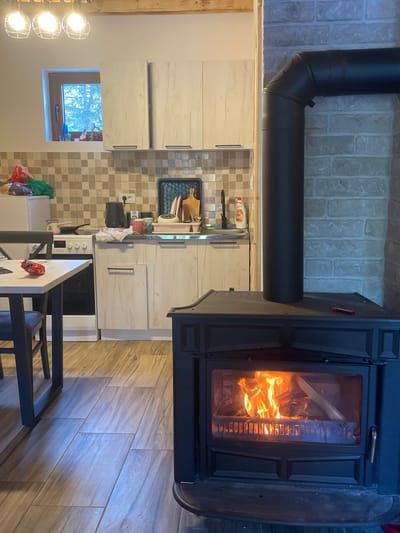 This image depicts an interior scene in Palež, Montenegro, featuring a rustic kitchen and an active wood-burning stove.
In the foreground, a dark, cast-iron wood-burning stove is prominently positioned on the right, with visible orange flames and burning wood through its glass door. A dark metal chimney pipe ascends from the stove, bending 90 degrees to the right and connecting to a light-colored brick wall. A small, red implement rests on the top surface of the stove. The floor consists of rectangular tiles with a light wood-grain pattern.
To the left of the stove, a light-colored table with dark metal legs is partially visible, with a dark chair tucked underneath. A crumpled red wrapper or bag lies on the table surface.
The background reveals a kitchen area. Light-toned wooden cabinetry, comprising both base and wall units with dark handles, lines the back wall. A stainless steel sink with a black faucet is present, flanked by a dish drying rack containing bowls and a cutting board, and a bottle of Fairy-brand dish soap. Various kitchen items, including a black kettle and a small white pot, are on the countertop. A beige and brown checkered tile backsplash covers the wall above the countertop. To the far left, a white refrigerator stands next to a white electric or gas stove with a frying pan on one burner. Above the upper cabinets, a small window offers a view of green foliage bathed in daylight. Exposed wooden beams are visible on the ceiling. No individuals are present in the scene.