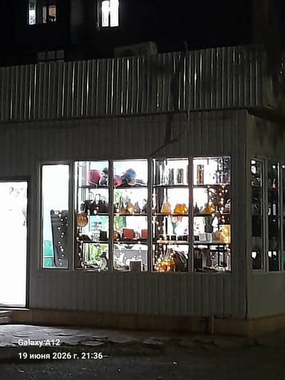 The image shows a storefront at night in the city of Kara-Köl, Kyrgyzstan. The storefront has large windows displaying various items, including what appear to be purses and decorative objects. Inside the store, shelves are filled with merchandise. A metal fence or corrugated siding runs along the top of the storefront. The lighting suggests it is nighttime, with some interior lights illuminating the store's contents. The bottom of the image contains text overlay indicating "Galaxy A12" and a date and time: "19 июня 2026 г. 21:36–".