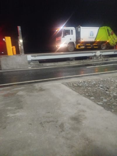 The image shows a white garbage truck with a green rear loader parked on a road at night. The truck has the logo "ТАЗАЛЫК" on its side. To the left of the truck are yellow bollards and a silver toll booth arm. The road appears to be wet, reflecting the lights from the truck and the surrounding environment. In the foreground, there is a concrete surface and a gravelly area. The background is dark, indicating it is nighttime. The location is identified as Түндүк айланып өтүүчү жол 84 га, Kyrgyzstan.