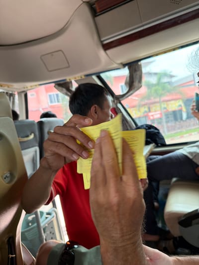 The image shows the interior of a bus, likely in the city of Phakhao, Laos. In the foreground, a person's hands are holding several yellow tickets. The hands are hairy and appear to be Caucasian. Behind the hands, a person wearing a red shirt is visible, holding more yellow tickets. They are sitting on a beige seat with dark grey upholstery. Other passengers are visible in the background, as are the windows of the bus, which offer a view of buildings and palm trees outside. The overall scene suggests a travel context.