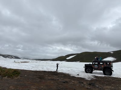 The image depicts a group of people in a rugged, snowy landscape, likely in or near Petropavlovsk-Kamchatskiy, Russia. The foreground is dominated by dark, rocky soil with patches of dry grass and scattered stones. Beyond this, a vast expanse of snow covers the ground, with some areas appearing more consolidated and others more churned or melted.

In the middle ground, a large, black, off-road vehicle with oversized tires is parked on the snow. The vehicle has a modified, rugged appearance, suggesting it's built for challenging terrain. Several people are visible on and around the vehicle. Three individuals are sitting on the roof rack of the vehicle, looking out at the surroundings. Another person is standing to the left of the vehicle, facing away from the camera and holding what appears to be a phone or camera up to take a picture.

The background features rolling, green hills, some of which are still capped with snow, indicating a high-altitude or early-season environment. The sky is overcast and gray, with thick clouds obscuring the sun, suggesting a cool, possibly damp, or wintry day. The overall mood of the image is one of adventure and exploration in a remote, natural setting. The contrast between the dark earth, the white snow, and the green hills creates a striking visual. There is a circular emblem with text on the side of the vehicle, but the text is not legible.