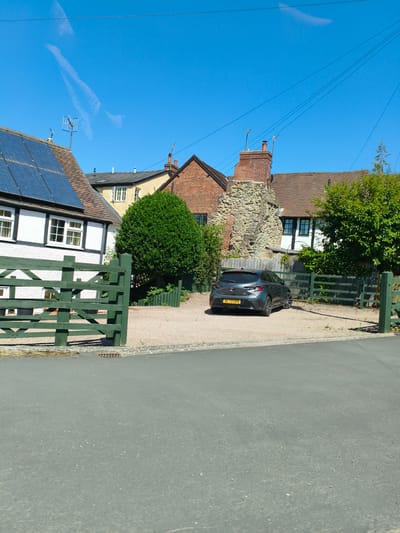 The image captures a view of a property with a dark grey hatchback car parked in the driveway, which is surfaced with gravel. The car's license plate reads "DL 73 GHA". A dark green wooden gate is visible in the foreground, partially obscuring the driveway. Behind the gate and car, there are buildings with tiled roofs, some featuring solar panels and chimneys. A stone ruin is also present. The scene is set against a clear blue sky. The location is Ludlow, United Kingdom.