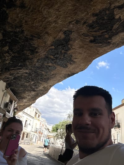 The image captures a selfie taken by a person in the foreground, who is holding a pink smartphone to take the photo. Another person is visible in the background, also looking at their phone. The scene appears to be outdoors, with a large, textured rock face dominating the left side of the frame. In the background, there are white buildings with balconies, characteristic of a Mediterranean or Spanish architectural style, suggesting the location is Setenil De Las Bodegas, Spain. Trees and foliage are also visible, indicating an outdoor setting possibly in a town or village. The sky is blue with scattered white clouds, suggesting daytime with fair weather. The overall mood seems casual and relaxed, with the people engaged in their devices. There is some faint text visible on a sign in the lower left corner of the image, but it is illegible. The perspective of the selfie is looking upwards towards the sky and the rock face, with the people and buildings further down.