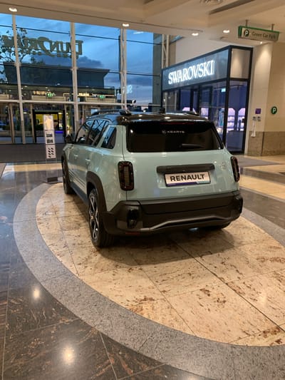 A light blue Renault car is parked indoors on a tiled floor of a shopping center. The back of the car faces the viewer. The tiles are a mix of light brown and gray shades and are arranged in a curved pattern. Through the glass windows behind the car, part of the shopping center can be seen with various store signs, including "SWAROVSKI" and "Milano", and other indistinct establishments. The store signs and general layout of the location indicate this is the Dundrum Town Centre, located in Dublin 16, Ireland. No people are visible in the immediate vicinity of the car. Signage indicating a Green Car Park is visible above the Swarovski store.