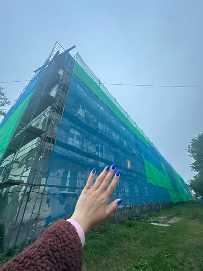 A low-angle, full shot shows a building covered in blue and green netting, indicating it is under renovation or repair. The building, which appears to be an apartment complex with multiple windows visible through the netting, is tall and angular, with scaffolding partially visible. In the foreground, a person's hand with bright blue painted fingernails is raised, obscuring part of the building. The hand is positioned in the lower right quadrant of the image. The environment around the building consists of a grassy area with some discolored patches, suggesting unkempt ground. The sky is overcast and a light color, contributing to a muted atmosphere. The scene is set in the city of Vilyuchinsk, Russia, a location known for its naval bases and proximity to volcanoes, though no specific landmarks in the image identify it as such beyond the provided context. The overall impression is one of ongoing construction work in a residential urban setting.