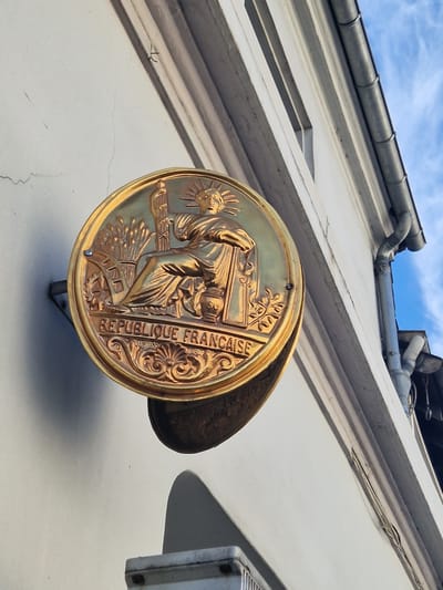 The image shows a close-up, low-angle shot of a circular golden seal affixed to the side of a building. The seal features a depiction of Marianne, a symbol of the French Republic, seated and holding a fasces, with a sprig of wheat and a cogwheel behind her. The inscription "REPUBLIQUE FRANCAISE" is visible on the lower portion of the seal. The building facade is painted white, and architectural elements like a gutter and downspout are visible. The sky is blue with some clouds. The scene is likely located in Antony, France, given the presence of the French Republic's seal.